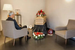 Homme en recueillement dans un salon funéraire devant un cercueil fleuri.
